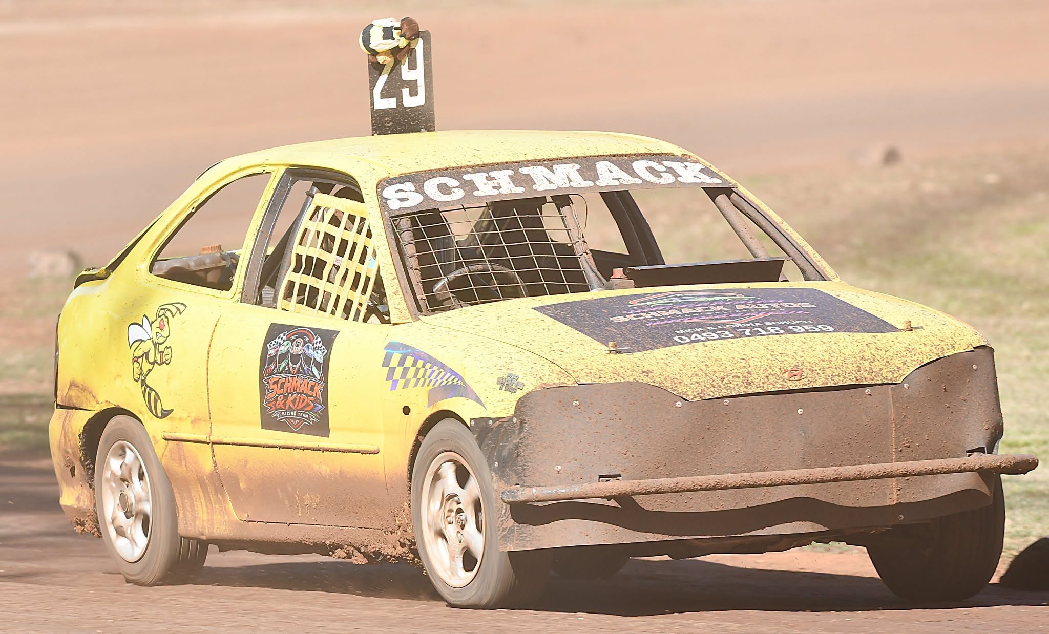 Yellow race car number 29 kicking up dust on a dirt track with a Schmack Autos bonnet sign
