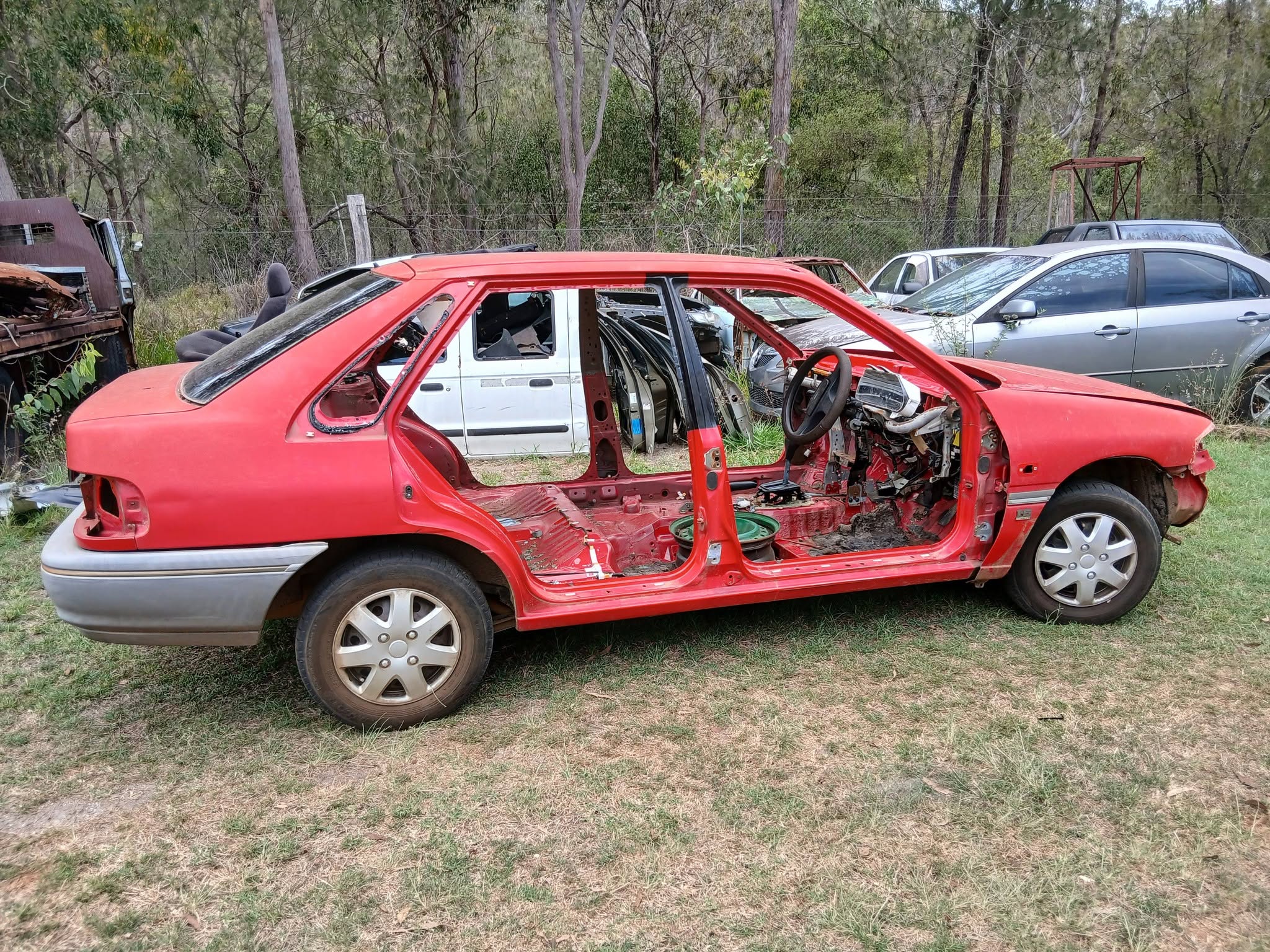Red car body with the interior removed, parked on grass beside the workshop yard